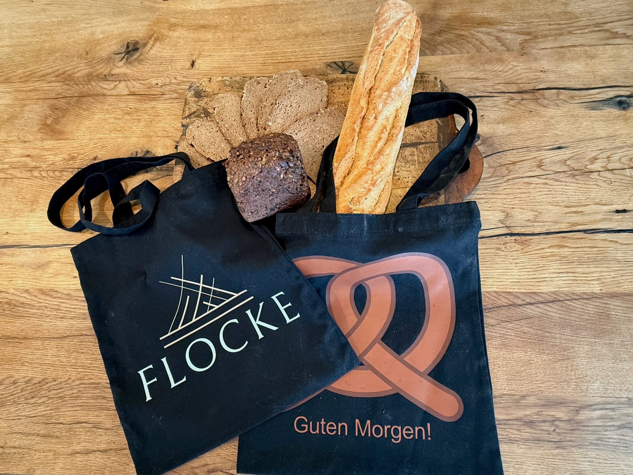 Zwei schwarze Tragetaschen auf einem Holztisch, eine mit Baguette und Roggenbrot, die andere mit geschnittenem Brot. Auf den Tüten steht „FLOCKE“ und „Guten Morgen!“.