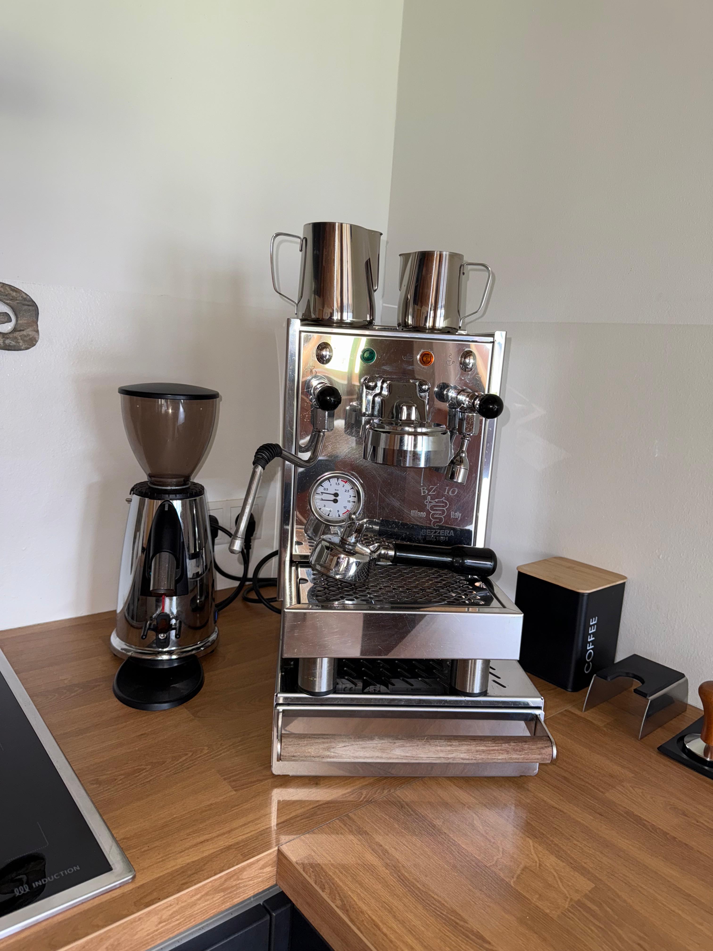 Eine Espressomaschine aus Edelstahl mit zwei Metalltassen obenauf, daneben eine Kaffeemühle auf einer hölzernen Arbeitsplatte.