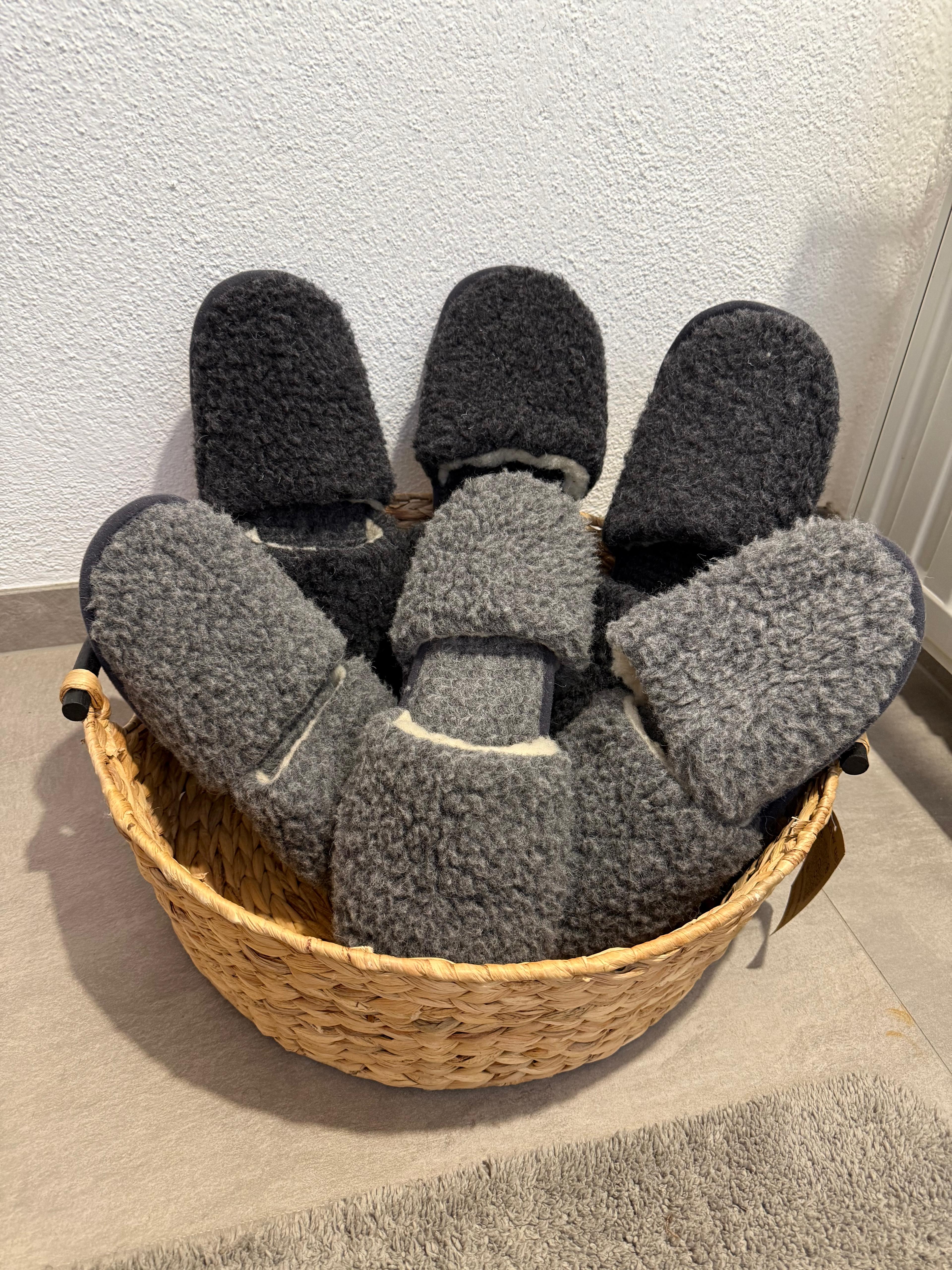 Ein geflochtener Korb, gefüllt mit mehreren Paaren grauer und schwarzer flauschiger Hausschuhe, steht auf einem gefliesten Boden vor einer weißen Wand.