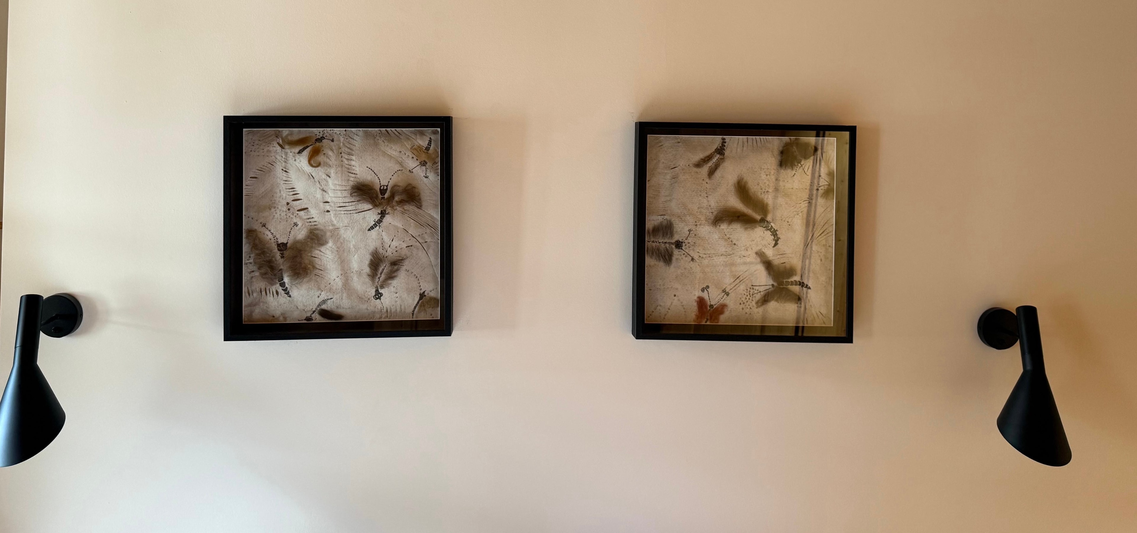 Zwei gerahmte abstrakte Kunstwerke in Erdtönen hängen an einer beigen Wand, flankiert von zwei schwarzen Wandlampen.